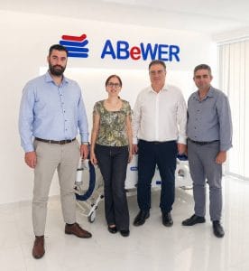 ABeWER leadership team at the company’s Cyprus headquarters, standing in front of the ABeWER logo with a multiTURN® 6 lateral turning mattress system on display behind them.