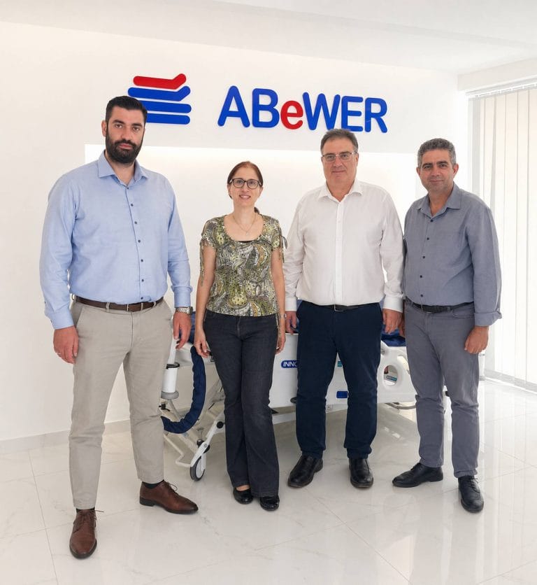 ABeWER leadership team at the company’s Cyprus headquarters, standing in front of the ABeWER logo with a multiTURN® 6 lateral turning mattress system on display behind them.