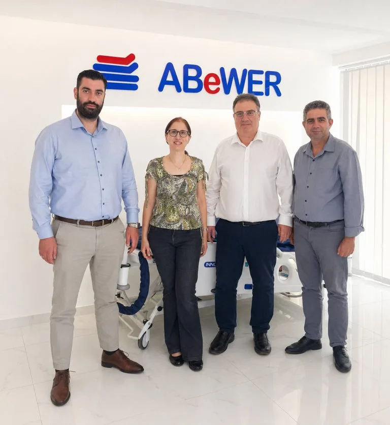 ABeWER leadership team at the company’s Cyprus headquarters, standing in front of the ABeWER logo with a multiTURN® 6 lateral turning mattress system on display behind them.